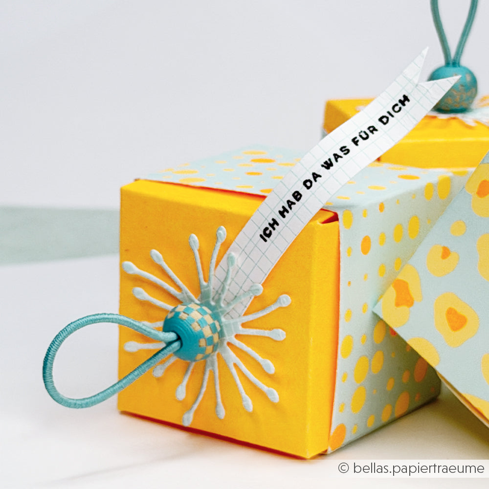 Gelbe Geschenkbox mit blauen Akzenten, einem Blumenmotiv aus leuchtendem Neonpapier und einem Anhänger mit der Aufschrift „ICH HAB DA WAS FÜR DICH“ – perfekt für kreative Papierprojekte oder um mit deinem Lieblings-Designpapier einen Farbtupfer zu setzen.