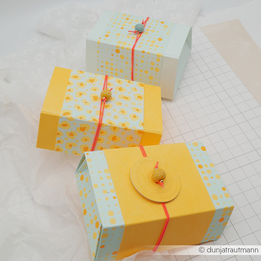 Drei farbenfrohe Geschenkboxen mit gelben und blauen Mustern, die mit einer roten Schnur zusammengebunden und oben mit kleinen Perlen verziert sind, wurden aus leuchtendem Neon-Papier und hochwertigem Designpapier gefertigt – perfekt für Ihre nächsten Papierprojekte.