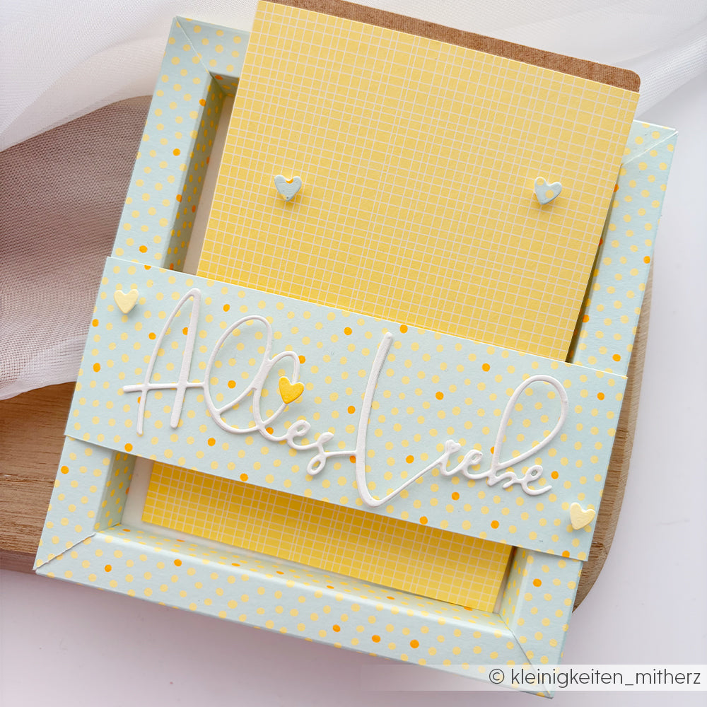 Gelbe Karte mit kleinen Herzen in einem blauen Rahmen mit Polka-Dots, gefertigt aus farbenfrohem Designpapier; der Text „Alles Liebe“ ist in weißer Schreibschrift aufgedruckt.