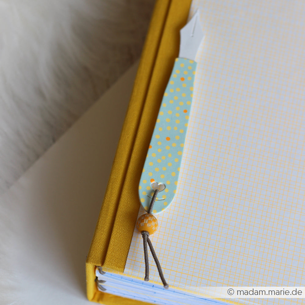 Gelbes Notizbuch mit einem Lesezeichen aus gepunktetem Designpapier „Sommer Special Streupünktchen“ und einer Perle, das auf einer weißen Unterlage liegt.