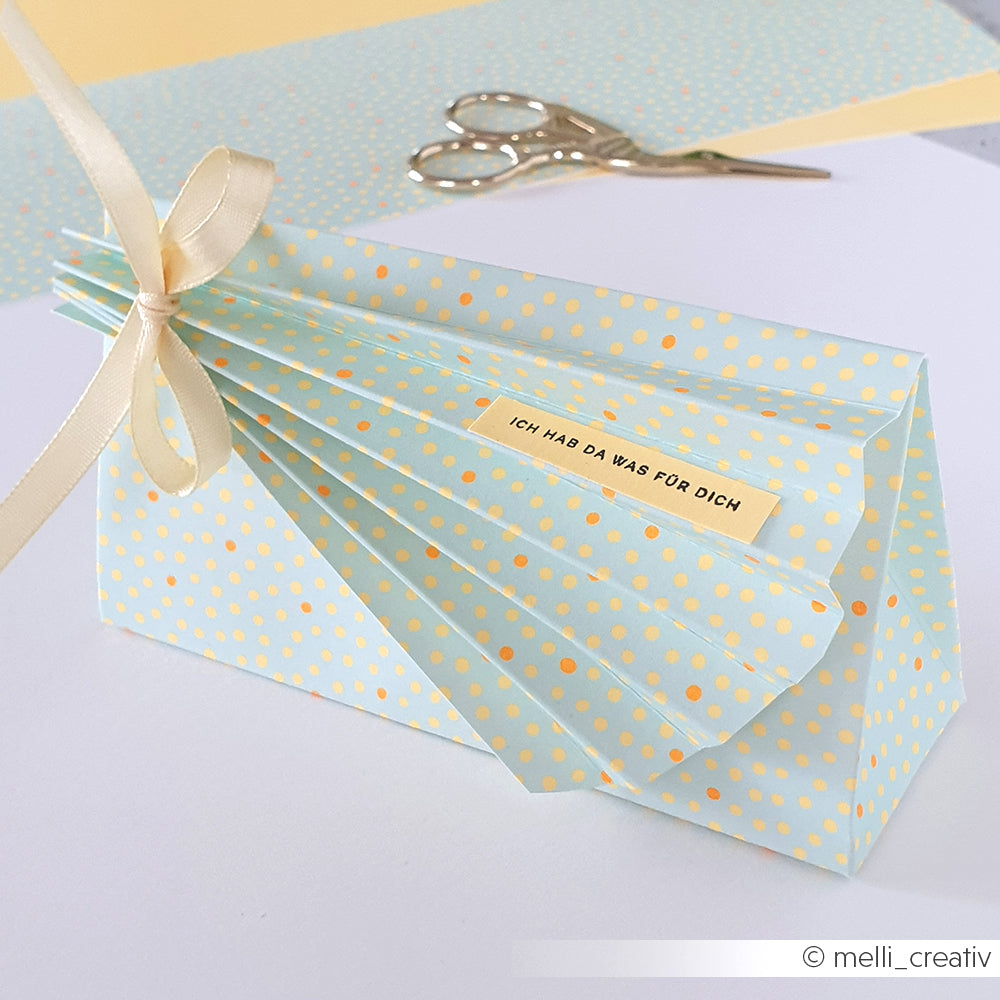 Eine blau-gelb gepunktete Origami-Geschenktüte aus farbenfrohem Designpapier, im Hintergrund sind ein Band, eine Schere und Bastelpapier zu sehen.