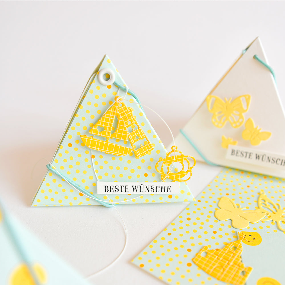 Eine dreieckige Karte mit gelben und weißen Punkten aus farbenfrohem Designpapier, verziert mit einem Segelboot und einem Anhänger mit der Aufschrift „Beste Wünsche“, präsentiert neben passenden Papierprojekten.