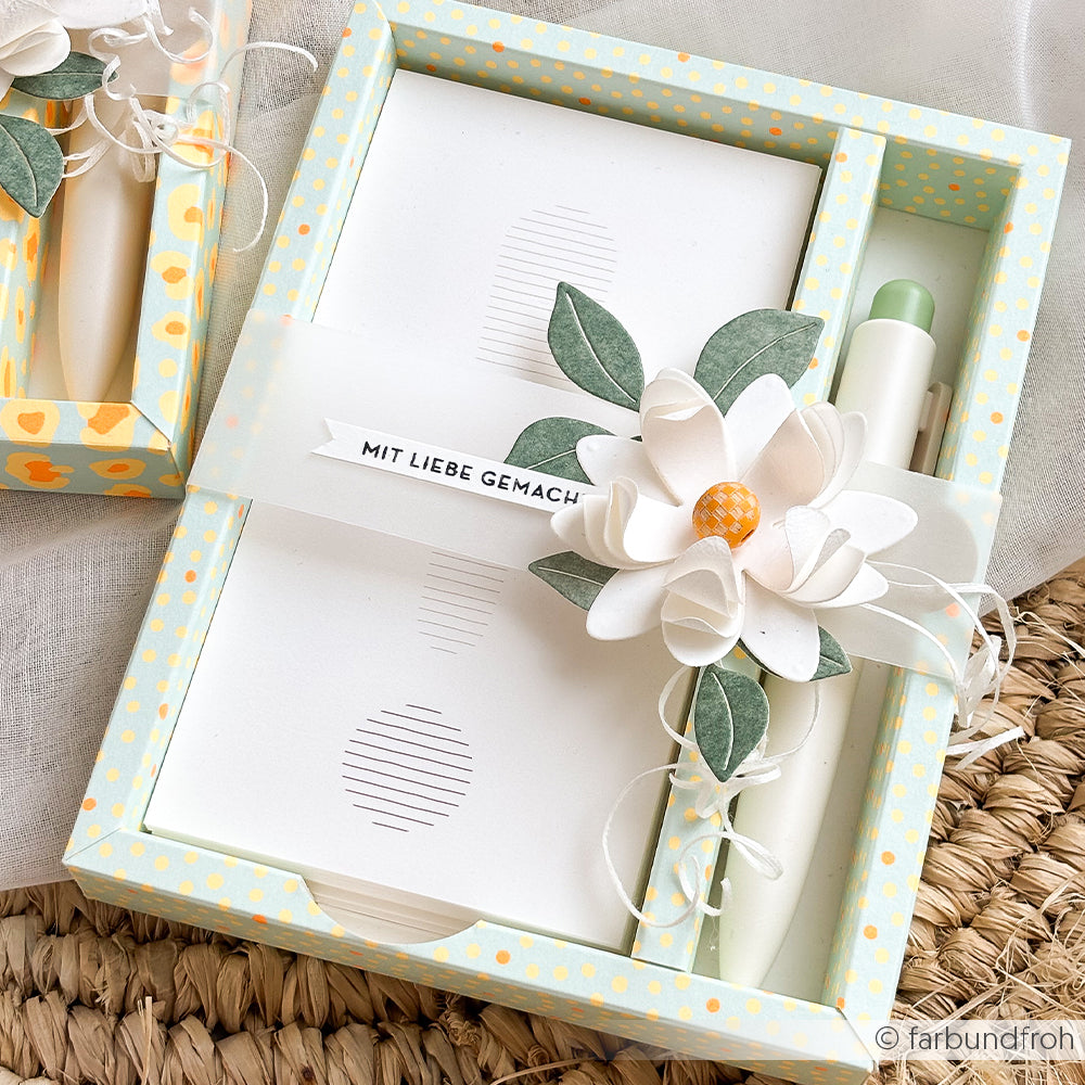 Geschenkbox mit einem Stift, einem Notizblock und einer weißen Papierblume aus Neon-Papier, verziert mit Blättern und einer Schleife.