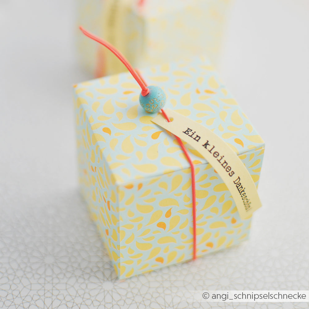 Kleine Geschenkbox aus Designpapier mit gelbem Muster, verziert mit einer roten Schnur, einer blauen Perle und einem Anhänger mit der Aufschrift „Ein kleines Dankeschön“. Ideal als Geschenk während der „Sommer Special Paisley“-Saison oder als fröhlicher Akzent mit leuchtenden Neon-Papier-Akzenten.