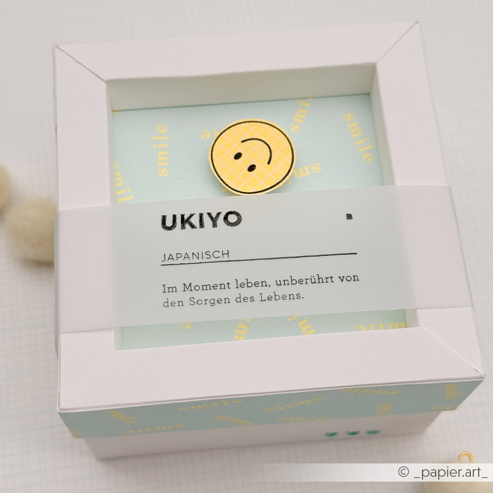 Weiße Geschenkbox mit einer Anstecknadel mit Smiley-Motiv und deutschem Text auf einem Etikett vor einem zartblauen, gemusterten Hintergrund – perfekt für Fans von Neon Papier oder alle, die ihren Papierprojekten eine fröhliche Note verleihen möchten.