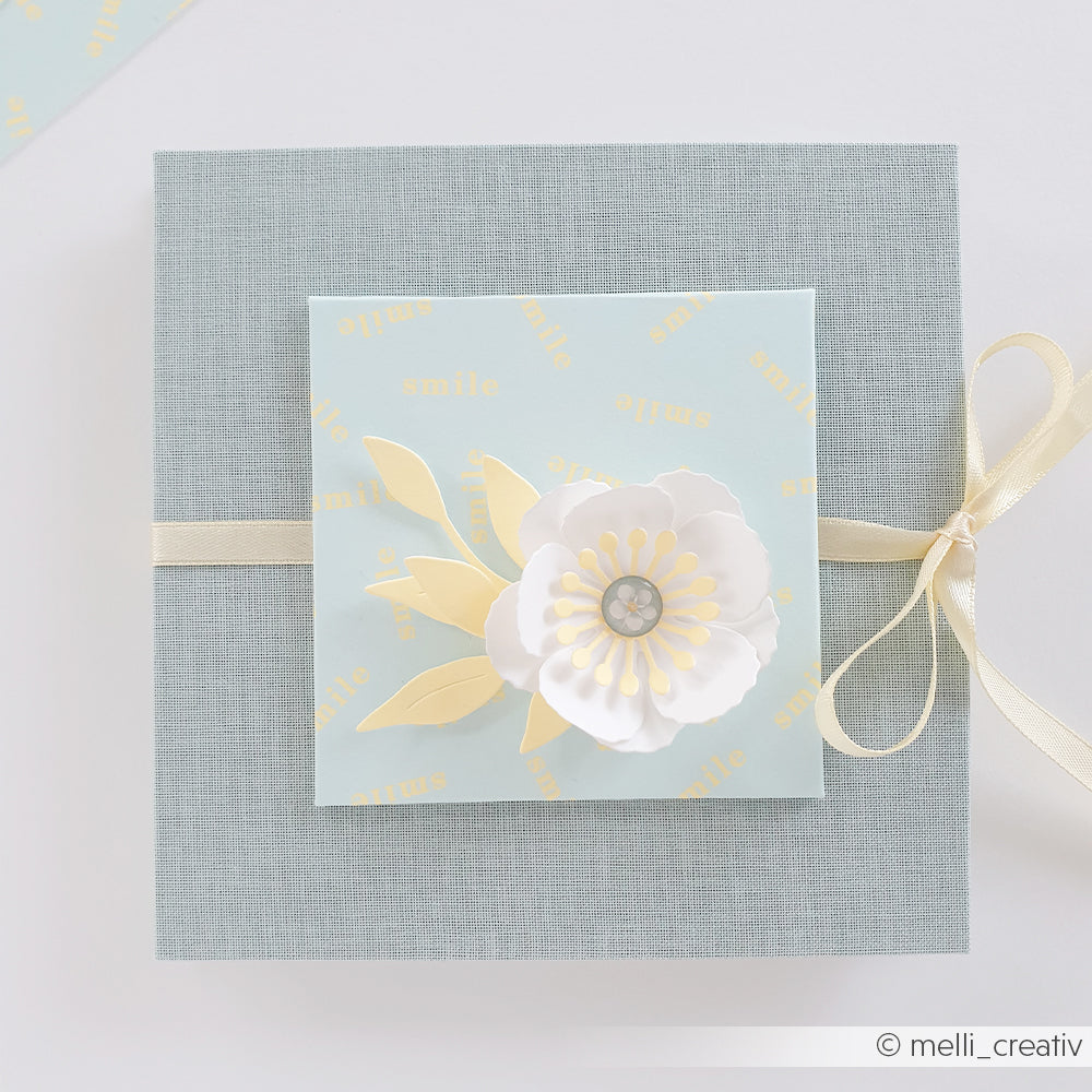Quadratische Geschenkbox mit Schleife, verziert mit einer Papierblume aus Designpapier und einem „Smile“-Schriftzug auf pastellblauem Hintergrund.