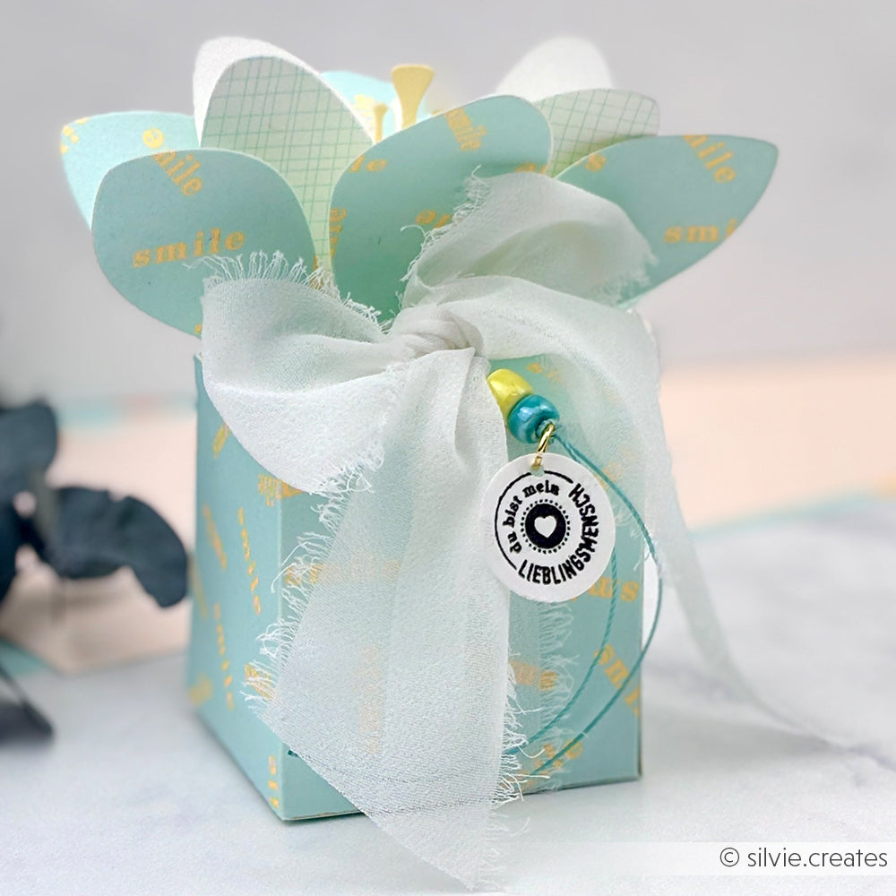 Eine blaue Geschenkbox mit einer großen weißen Schleife, herzförmigen Blütenblättern aus Neonpapier und einem runden Anhänger, der am Band hängt – perfekt, um Ihren Papierprojekten eine kreative Note zu verleihen oder als Inspiration für einzigartige Designpapier-Ideen.