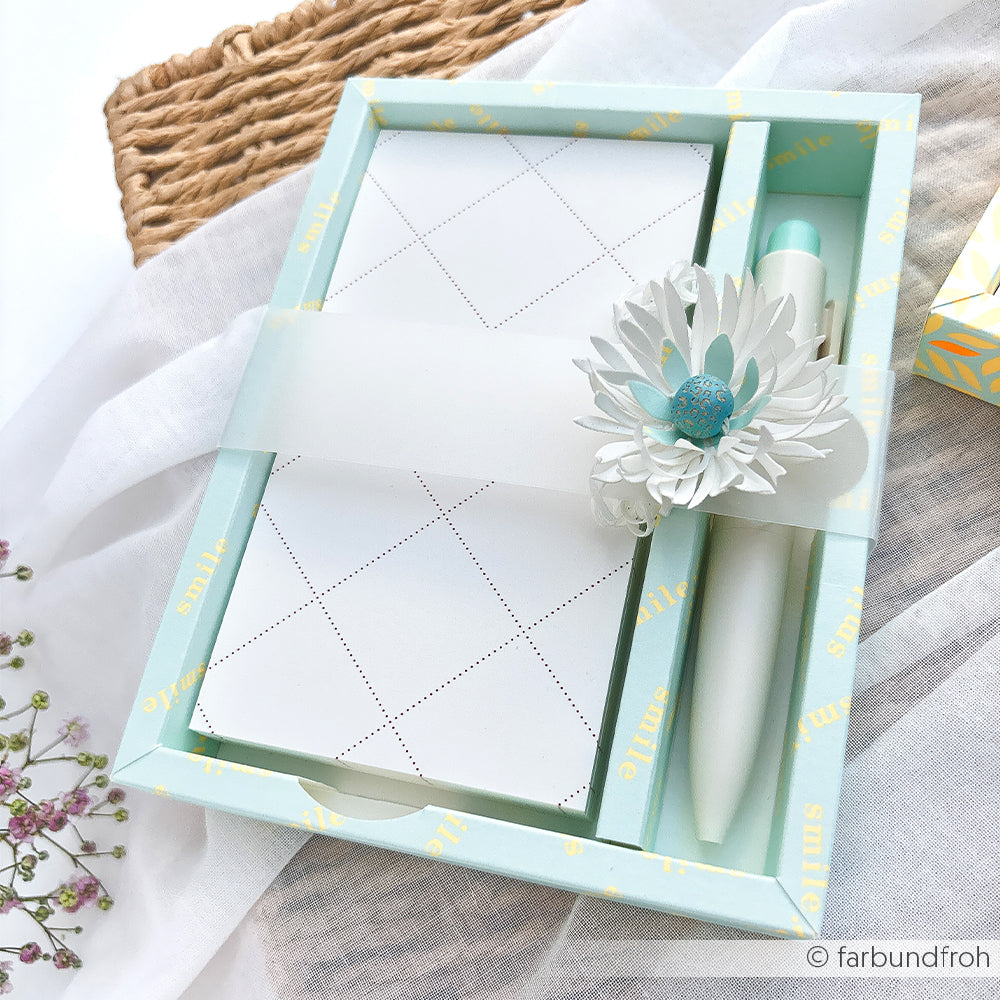Geschenkbox mit einem Stift, einem Notizblock aus stilvollem Designpapier und einer weißen Blumendekoration auf weißem Hintergrund.