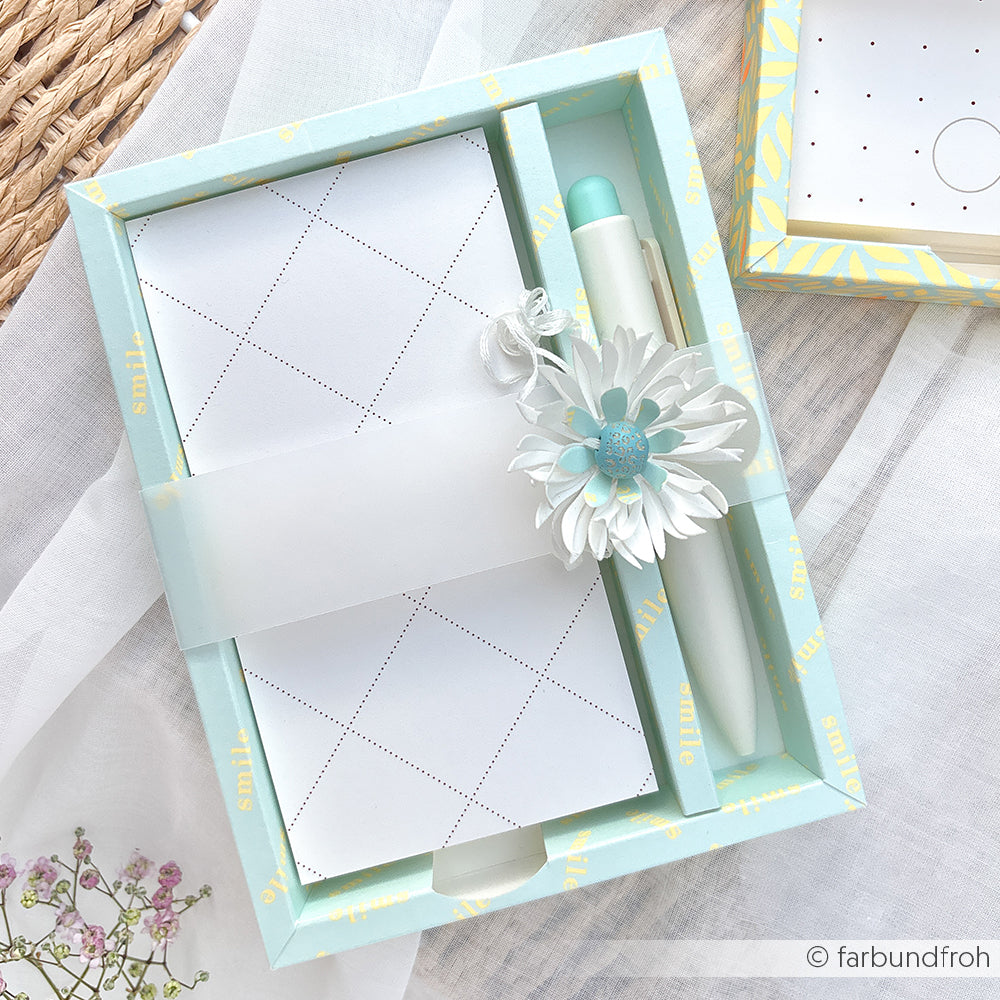 Ein Schreibwaren-Set in Pastelltönen mit einem Notizblock aus hochwertigem Designpapier, einem Stift mit Blumenanhänger und einer zarten Verpackung auf einer geflochtenen Unterlage.