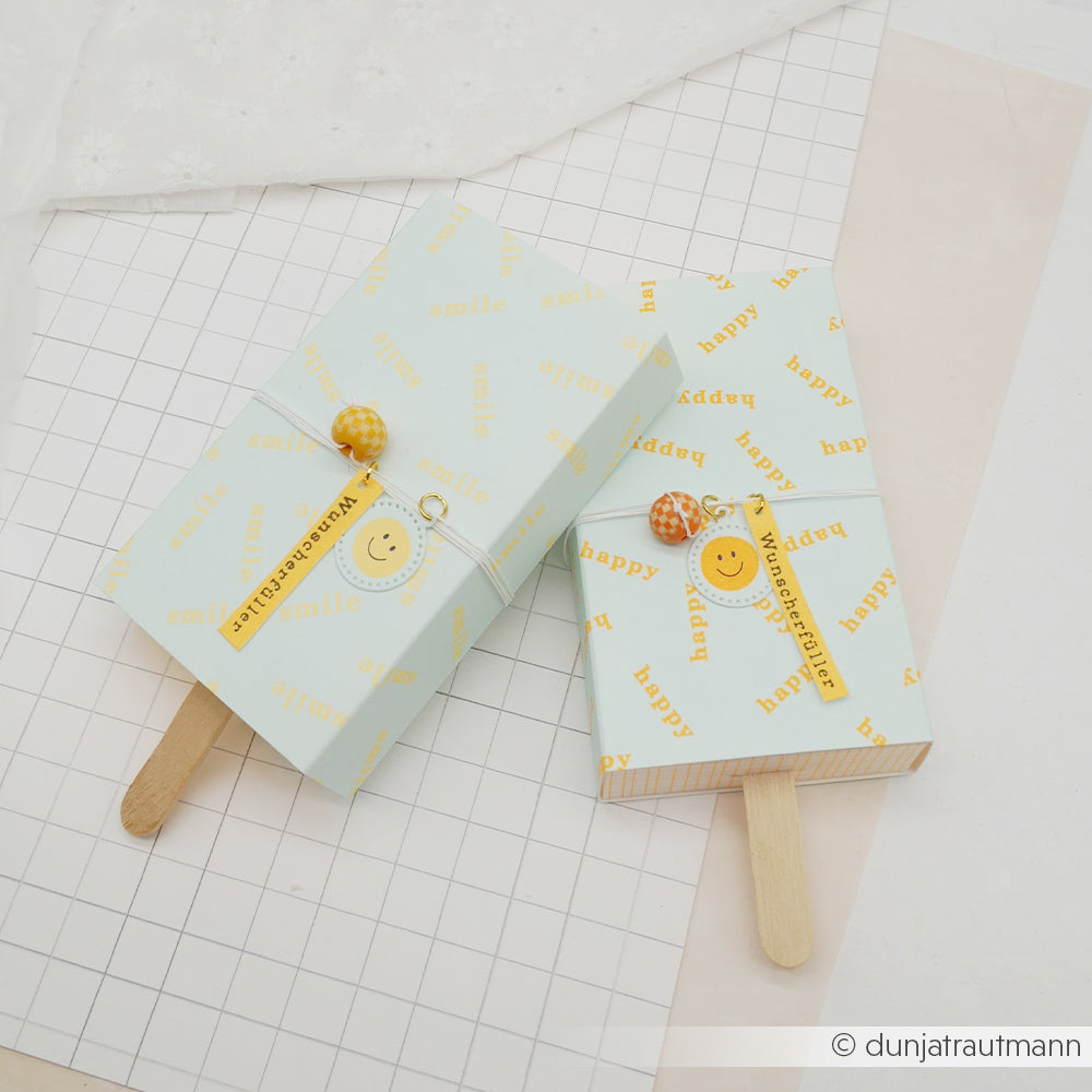 Zwei pastellfarbene Geschenkboxen mit den Aufschriften „happy“ und „smile“, verziert mit gelben Anhängern und Lutscherstielen, gefertigt aus hochwertigem Designpapier und ideal für kreative Papierprojekte.