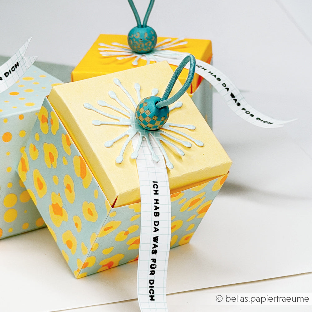 Farbenfrohe Geschenkboxen mit gemusterten Motiven aus Neonpapier und Designpapier, verziert mit Perlen und Anhängern aus Band, auf denen „Ich hab da was für dich“ steht.