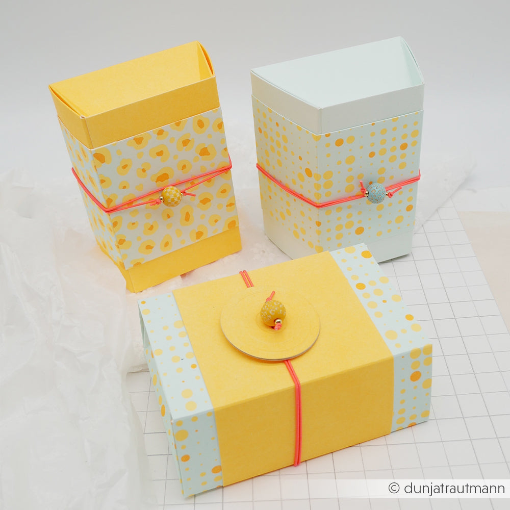 Drei pastellfarbene Geschenkboxen mit gelben Mustern, verziert mit einer rosa Schnur und kleinen Fisch-Anhängern. Jede Box ist aus hochwertigem Designpapier gefertigt und verleiht dem verspielten Design einen Hauch von Eleganz.