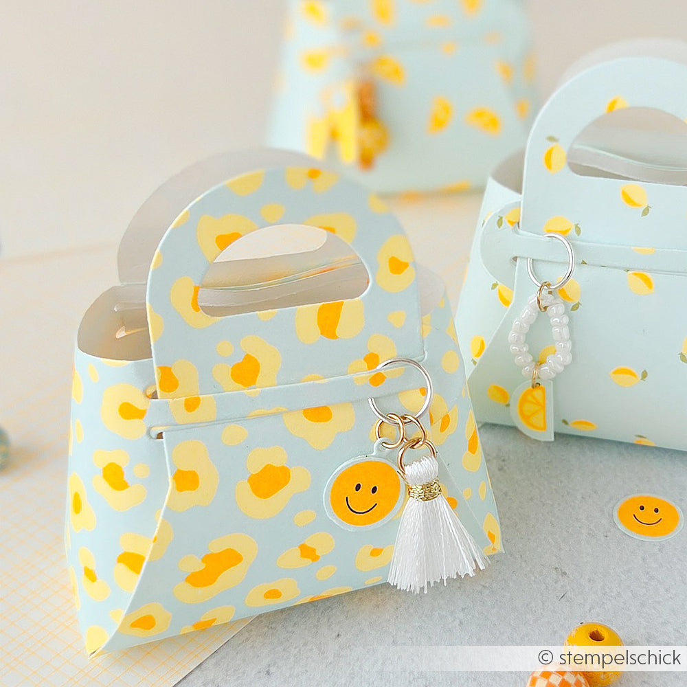 Kleine pastellblaue Geschenktüten mit gelben Punkten, einem Smiley-Aufkleber und einem weißen Schlüsselanhänger mit Quaste – jede einzelne aus hochwertigem Designpapier gefertigt, was ihnen das gewisse Etwas verleiht.