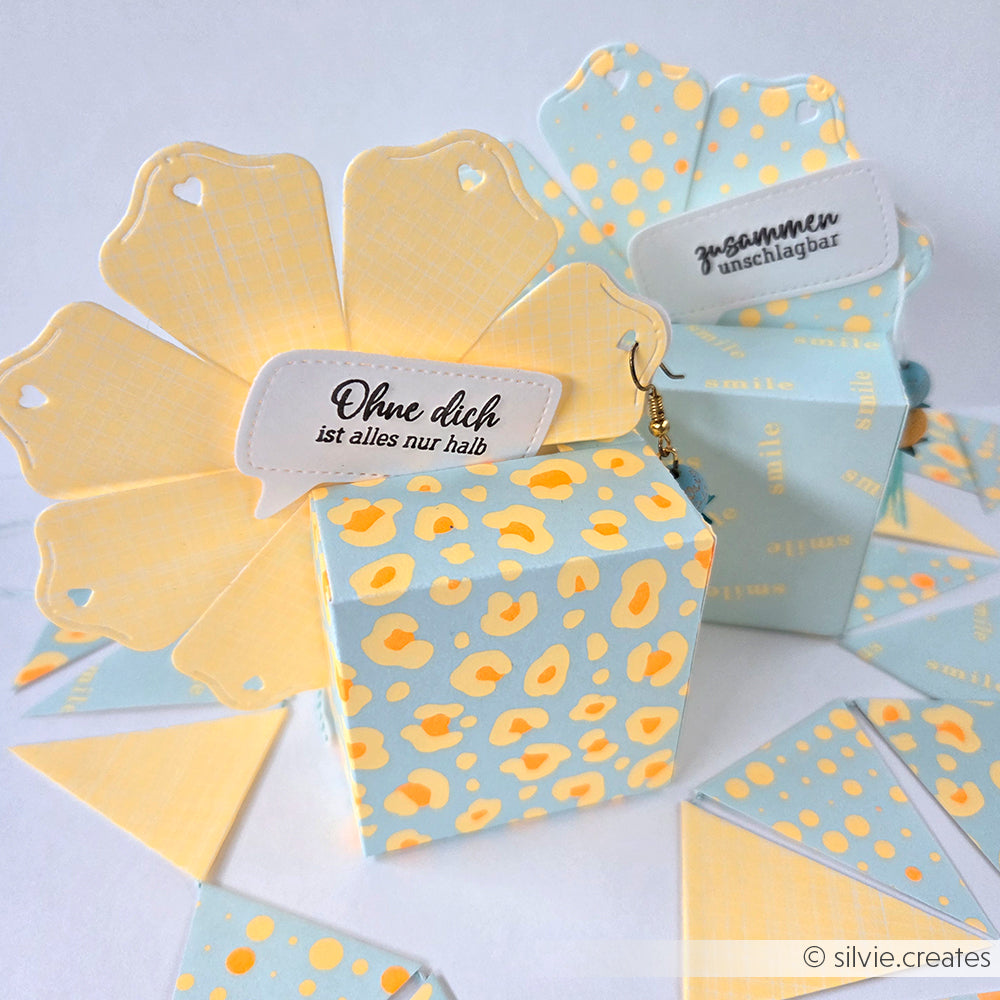 Zwei pastellfarbene Geschenkboxen mit deutschen Aufschriften, eine offen und eine geschlossen, verziert mit gelben und blauen Mustern aus farbenfrohem Designpapier, die für einen stilvollen Akzent sorgen.