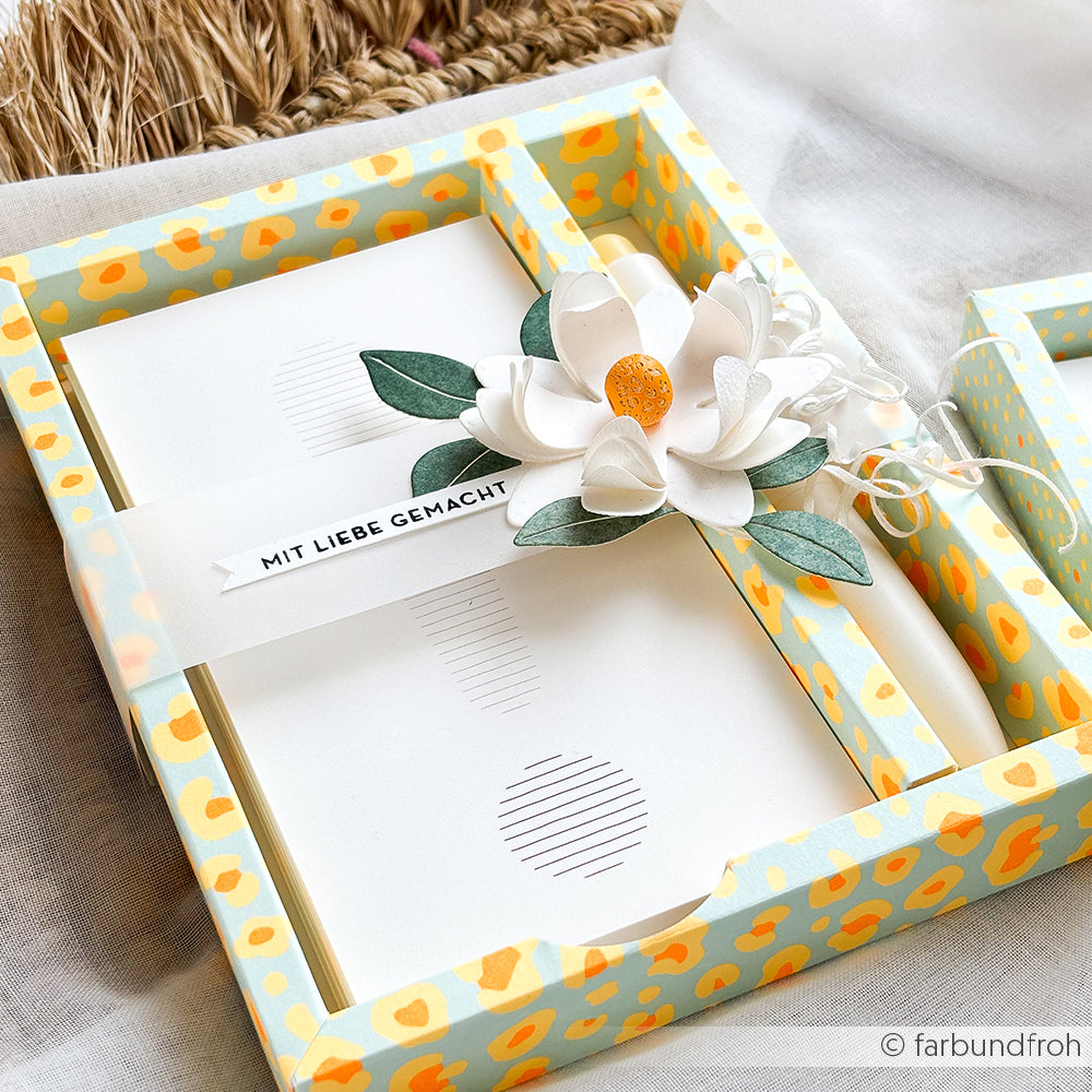 Eine mit Blumen verzierte Geschenkbox mit Briefpapier, einem Anhänger mit der Aufschrift „MIT LIEBE GEMACHT“ und einem gemusterten gelben Motiv aus leuchtendem Neonpapier.