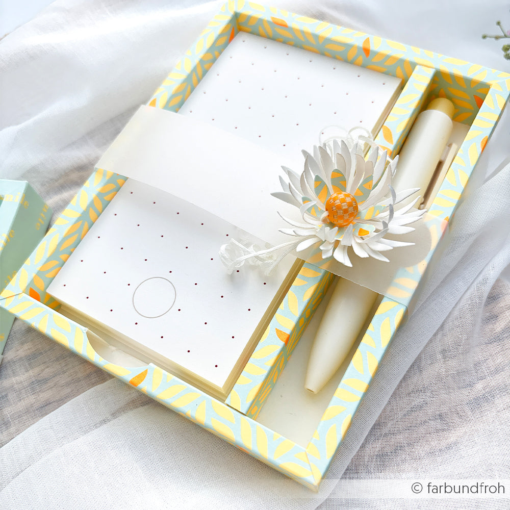 Geschenkset mit Notizblock und Stift in einer gelb gemusterten Schachtel im „Neon Papier“-Design, verziert mit einer weißen Papierblume und einer orangefarbenen Perle – ein perfektes Sommer-Special.