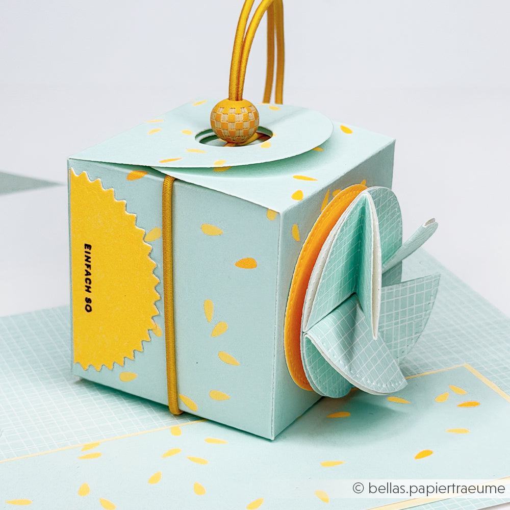 Eine pastellblau-gelb verzierte Geschenkbox aus zartem Designpapier mit einem Schnurgriff und runden Blütenblatt-Verzierungen.