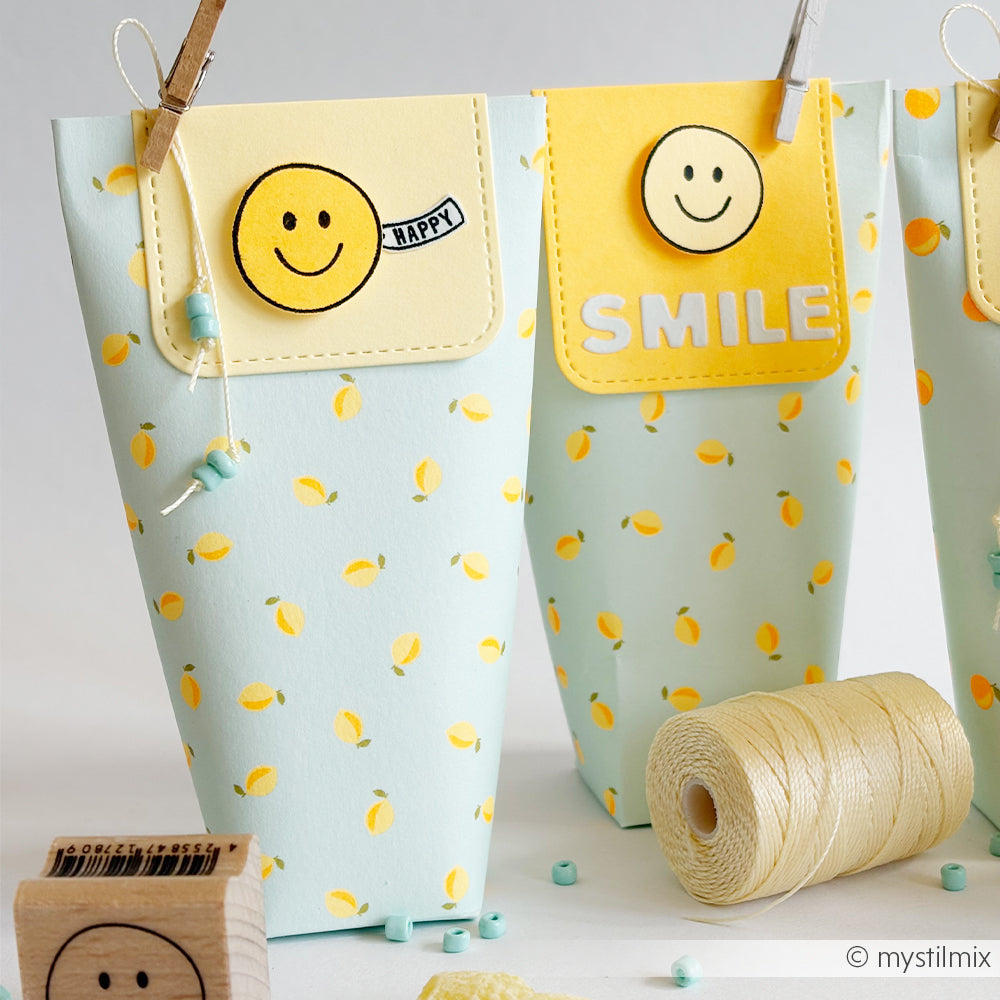 Auf einem Tisch liegen zwei Geschenktüten mit Zitronenmotiv aus Designpapier, verziert mit fröhlichen gelben Smileys und den Aufschriften „HAPPY“ und „SMILE“ – perfekte Papierprojekte für Ihre „Sommer Special Zitronen“-Feier.