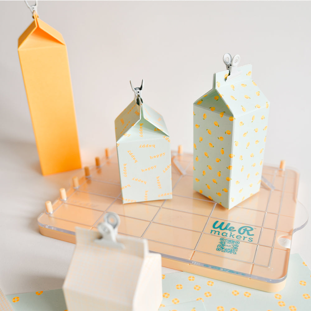 Pastellfarbene Papierkartons mit Mustern, darunter „Designpapier Neon“ und „Papier für Grußkarten“, stehen auf einer Bastelunterlage mit der Aufschrift „We R makers“.