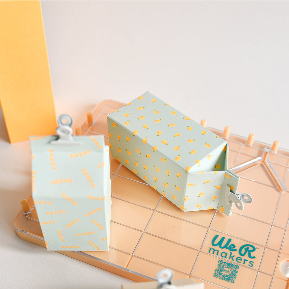 Zwei pastellfarbene Papierschachteln mit Mustern und Klammern stehen auf einer pfirsichfarbenen Bastelunterlage mit der Aufschrift „We R makers“ – ideal, um Ihr „Neon Papier“ oder das „Designpapier Summer Special Zitronen“ in kreativen Papierprojekten in Szene zu setzen.