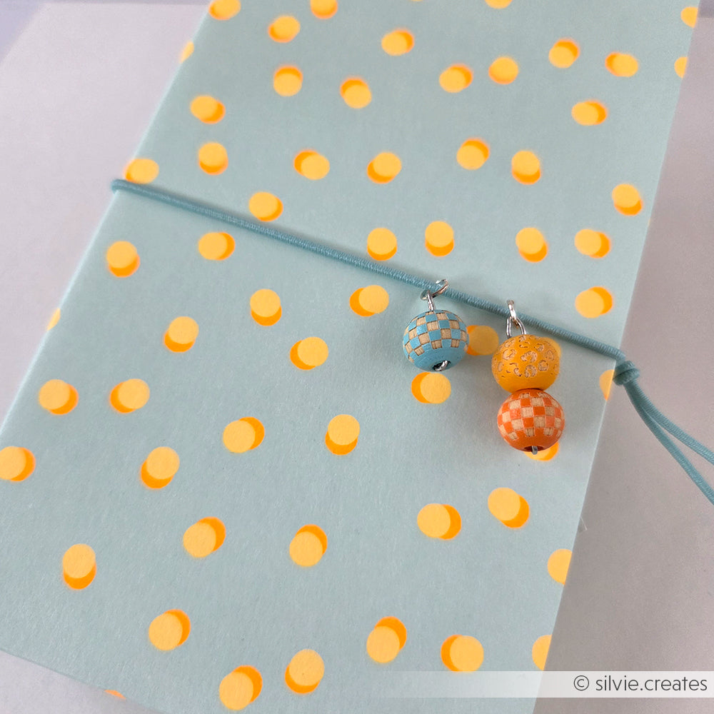 Hellblaues Notizbuch mit gelben Punkten, verziert mit drei bunten Perlenanhängern an einem Gummiband – perfekt für Papierprojekte oder um mit Neonpapier und einzigartigen Designpapier-Akzenten eine verspielte Note zu setzen.