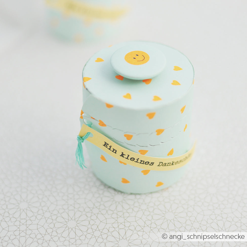 Hellblaue, runde Geschenkbox mit gelben Herzen, einem Smiley auf der Oberseite und einem Anhänger mit der Aufschrift „Ein kleines Dankeschön“. Ideal in Kombination mit Grußkarten oder zum Einpacken in auffälliges Neonpapier, um eine wirklich unvergessliche Präsentation zu schaffen.