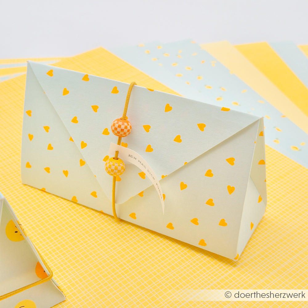 Ein Papierumschlag mit gelben Herzen, mit einer Schnur und Perlen verschnürt, auf schickem „Neon Papier“-Designpapier platziert – perfekt für einen sommerlichen „Herzen“-Akzent.