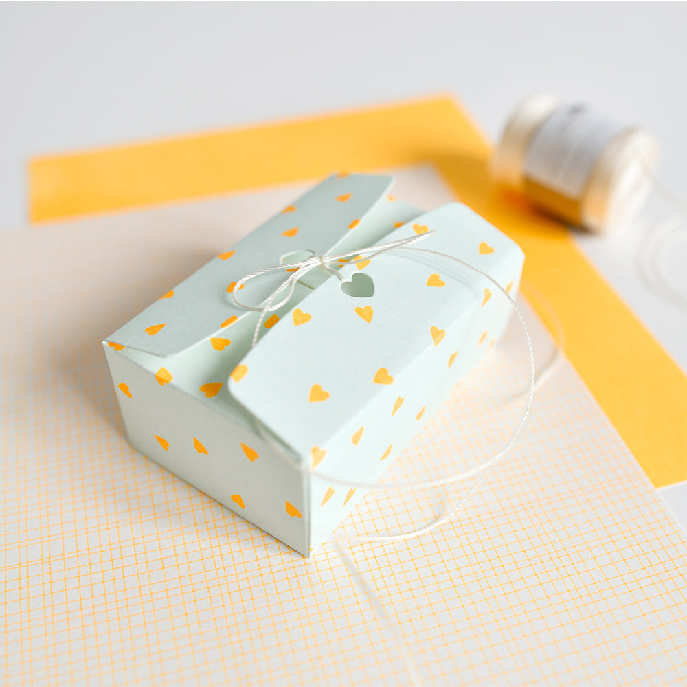 Eine hellblaue Geschenkbox mit gelben Herzen, die mit einer Schnur zusammengebunden ist, steht auf gelb-weiß gemustertem Designpapier – ideal für kreative Papierprojekte.