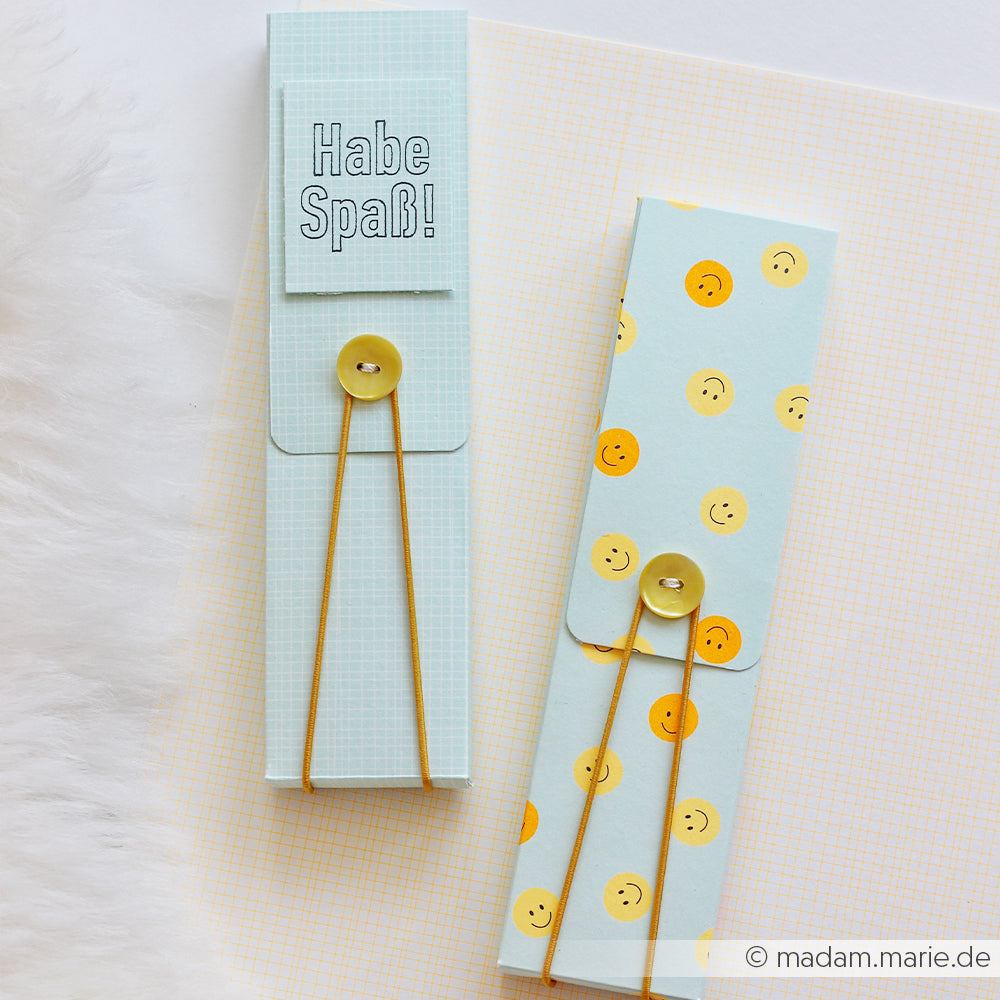 Zwei pastellblaue Papierumschläge mit Knöpfen, gefertigt aus hochwertigem Designpapier – auf dem einen steht „Habe Spaß!“, auf dem anderen sind fröhliche gelbe Smileys abgebildet.