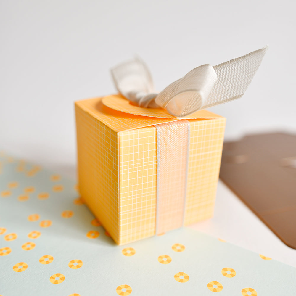 Eine gelbe Geschenkbox mit einer weißen Schleife steht auf einem hellblauen Untergrund mit gelben Punkten und schafft so eine verspielte Szene, die an farbenfrohes Designpapier aus der Buchbinderei oder sogar an bunte Akzente aus Neonpapier erinnert.