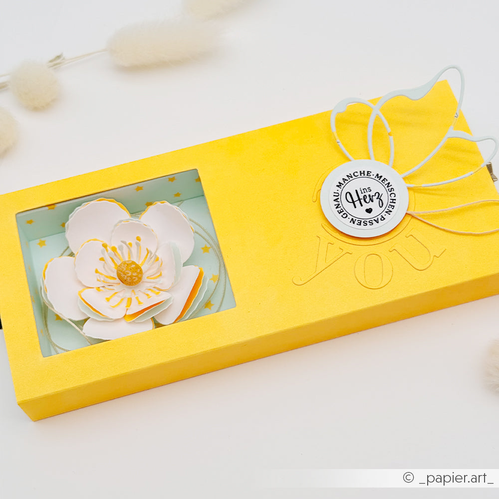 Gelbe Geschenkbox mit einer weißen Papierblume und einem runden Anhänger, verziert mit zarten weißen Akzenten – perfekt zum Präsentieren von Grußkarten oder als elegante Dekoration neben Ihrem Lieblings-Designpapier.