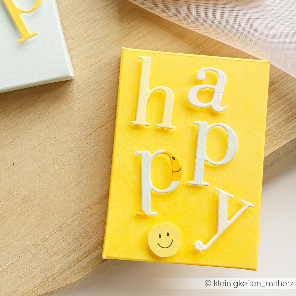 Gelbe Karte mit dem Wort „happy“ in weißen Buchstaben und Smiley-Aufklebern auf hellem Holzhintergrund – gefertigt aus leuchtendem Neon-Papier in Sonnengelb für einen strahlenden und fröhlichen Akzent.