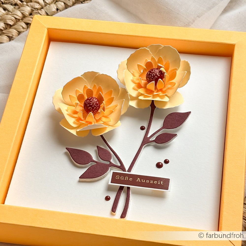 Schattenkastenrahmen mit zwei mehrschichtigen Papierblumen aus dem Mimi Summer Special „Leuchtend Sonnengelb“-Designpapier und Neonpapier, mit dem deutschen Schriftzug „Süße Auszeit“ auf weißem Hintergrund.