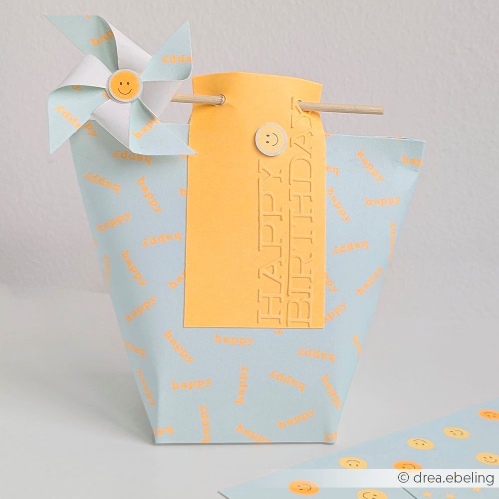 Hellblaue Geschenktüte mit Geburtstagsgruß, einem gelben „HAPPY BIRTHDAY“-Anhänger aus Mimi Summer Special Leuchtend Orange Neon Papier und einer Windrad-Verzierung aus farbenfrohem Designpapier.