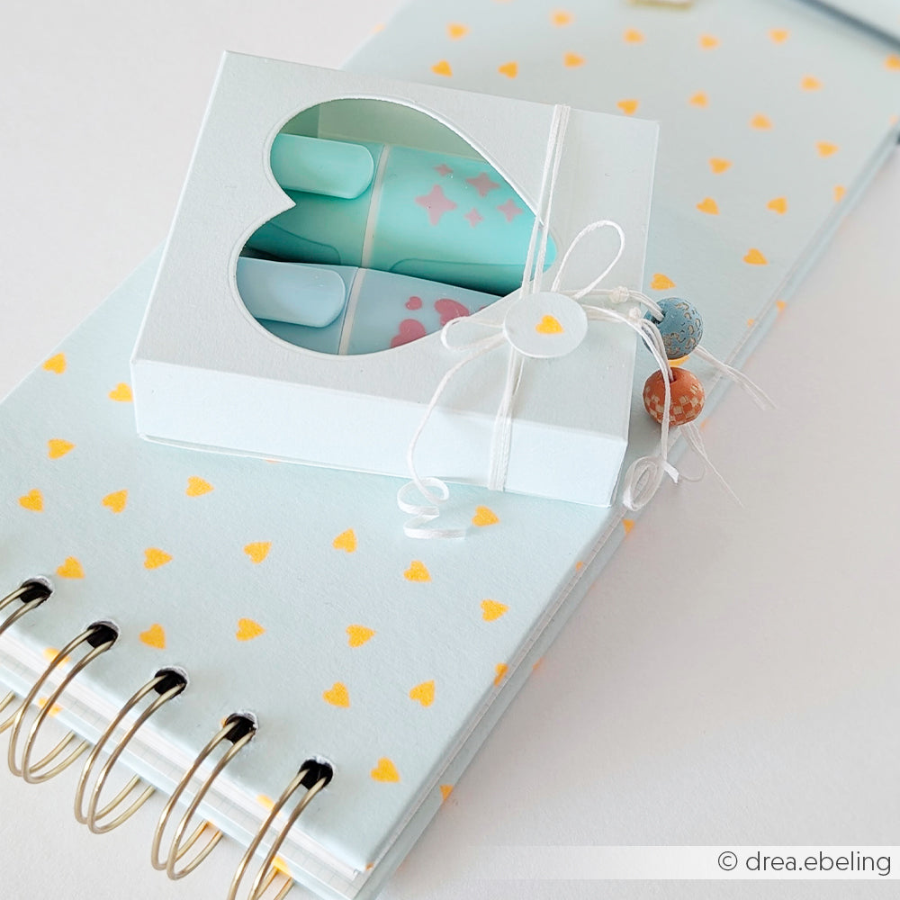 Hellblaues Notizbuch mit gelben Herzen aus Neon Papier Designpapier, verziert mit einer kleinen, mit einer Schnur verschnürten Geschenkbox auf der Oberseite – ein perfektes Detail aus der Mimi Summer Special „Leuchtend Aqua“-Kollektion.