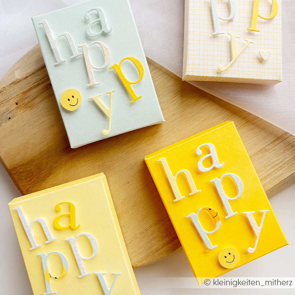 Vier pastellfarbene und gelbe Geschenkboxen aus Neon-Papier und Designpapier, jeweils mit dem Schriftzug „happy“ in Reliefbuchstaben und verziert mit kleinen Smiley-Knöpfen.