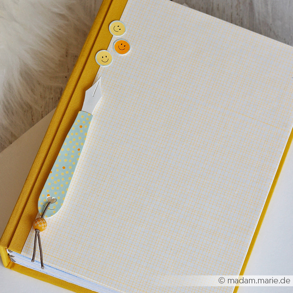 Ein gelbes Notizbuch mit kariertem Papier aus Designpapier „Sonnengelb“, Smiley-Aufkleber und ein blauer, gepunkteter Füllfederhalter auf weißem Hintergrund.