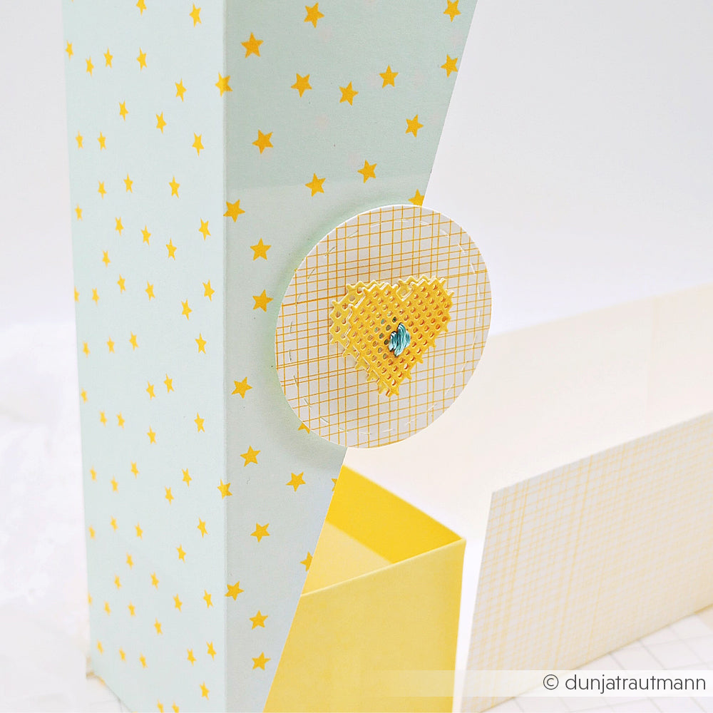 Hohe Geschenkbox mit Sternenmuster aus Neon-Papier, verziert mit einem runden, gelben Kreuzstich-Herzmotiv und Gitterdetails – perfekt für stilvolle Papierprojekte.