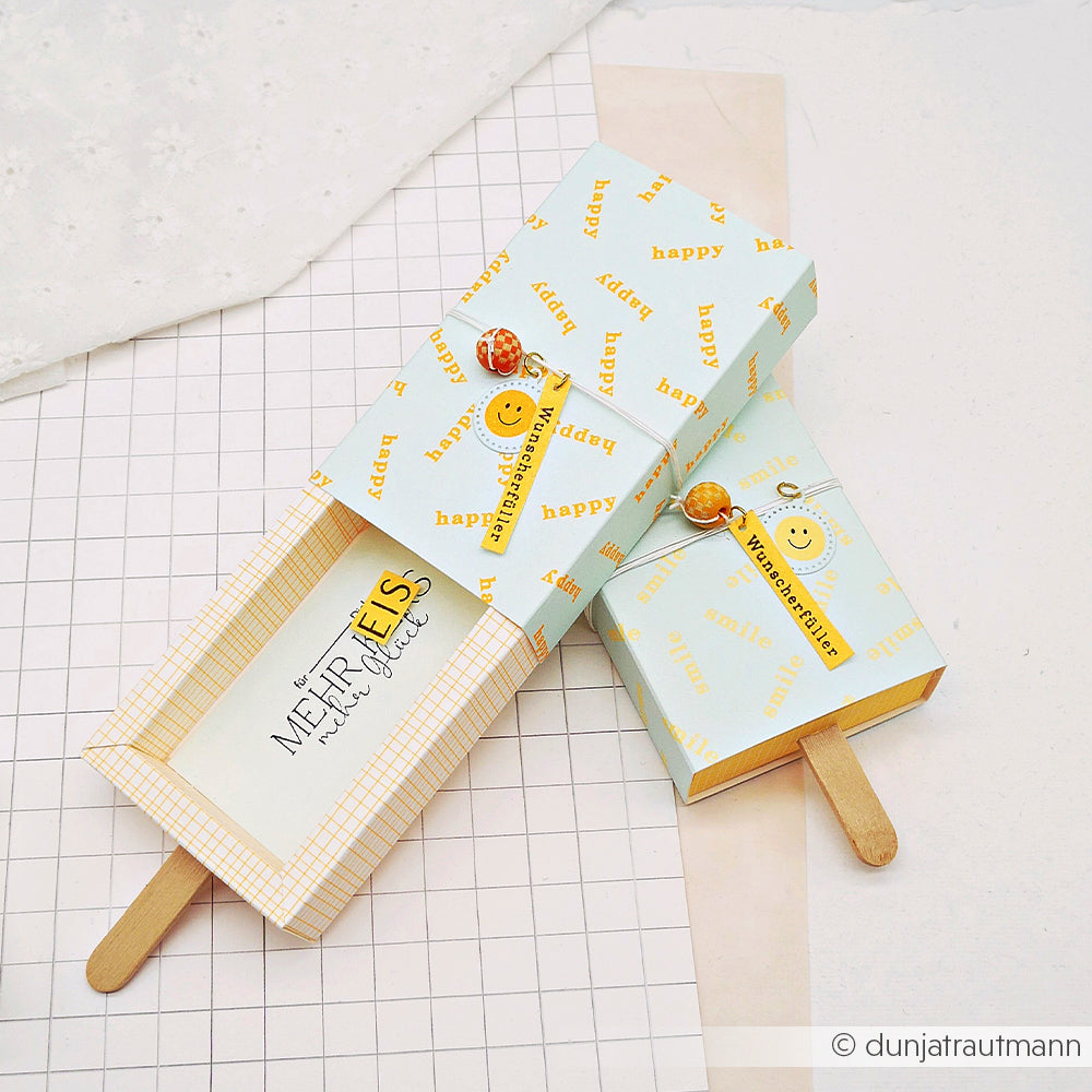 Zwei pastellfarbene Geschenkboxen in Eis am Stiel-Form mit Stielen, gefertigt aus farbenfrohem Designpapier und verziert mit fröhlichen Sprüchen und Smileys.