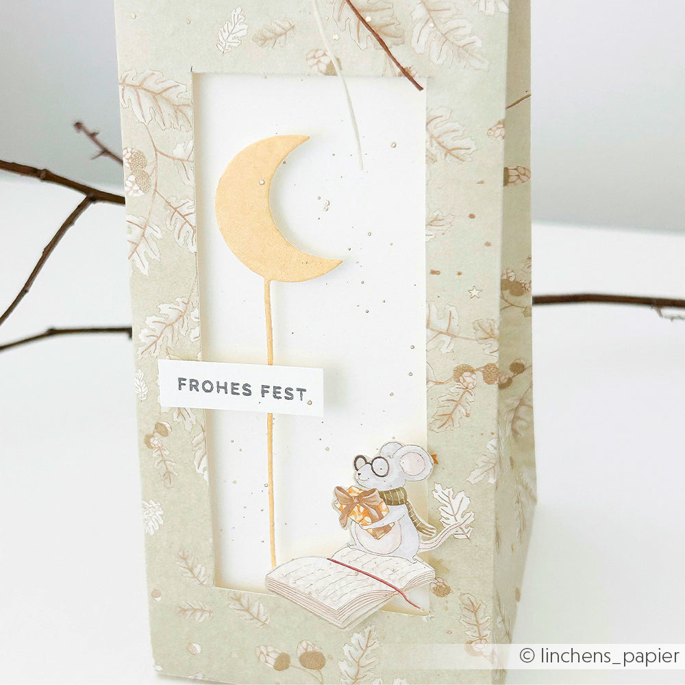 Eine dekorative Weihnachtstüte aus Designpapier mit einer Maus, die ein Geschenk trägt, einem Buch, einem Mond und der Aufschrift FROHES FEST.