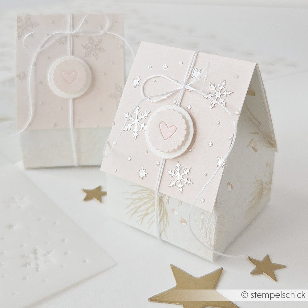 Zwei hellrosa, hausförmige Geschenkboxen aus Designpapier mit Schneeflocken, Schnur und einem Herzsiegel.