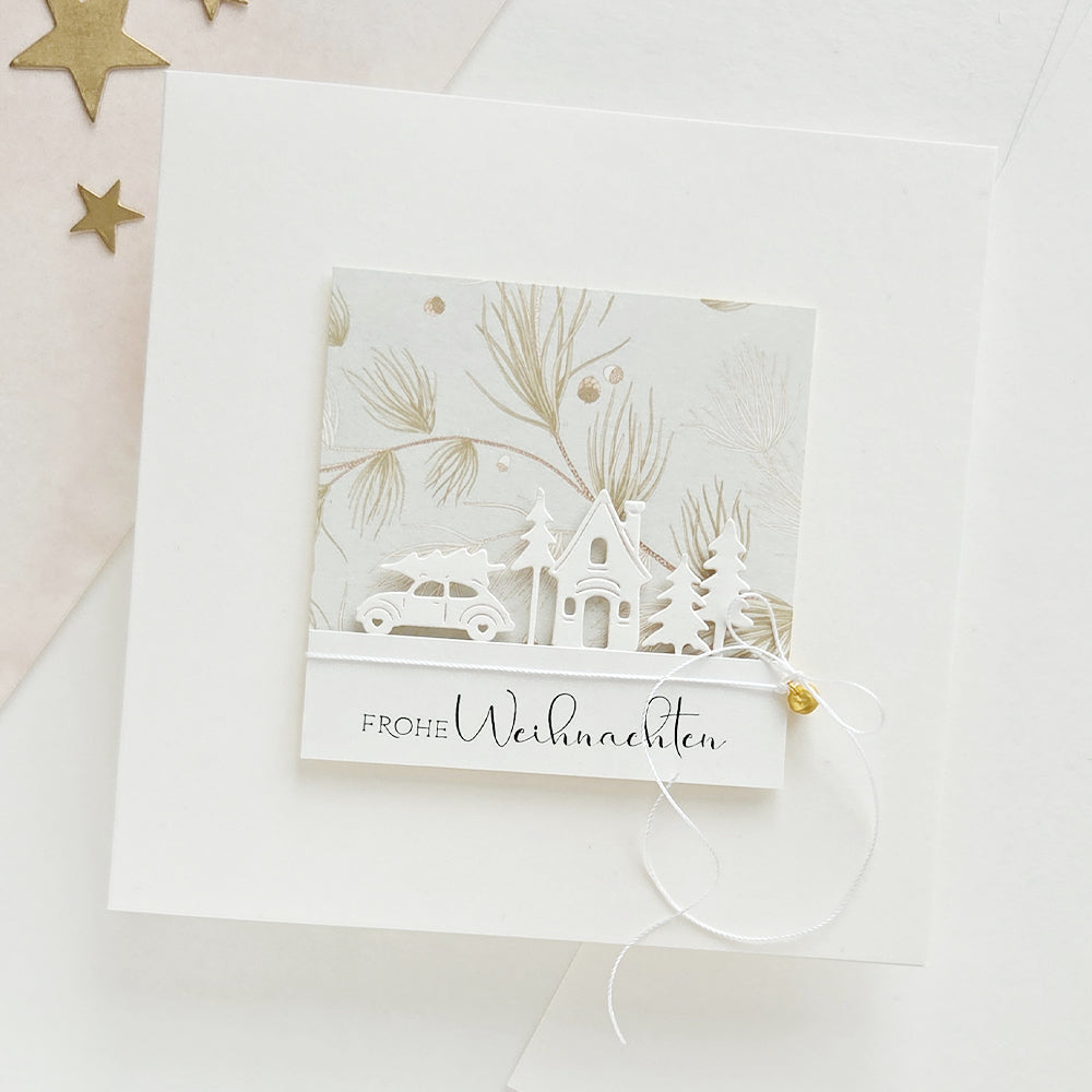 Quadratische Weihnachtskarte mit einem Motiv aus einem Haus, Bäumen und einem Auto auf Designpapier, mit der Aufschrift Frohe Weihnachten.