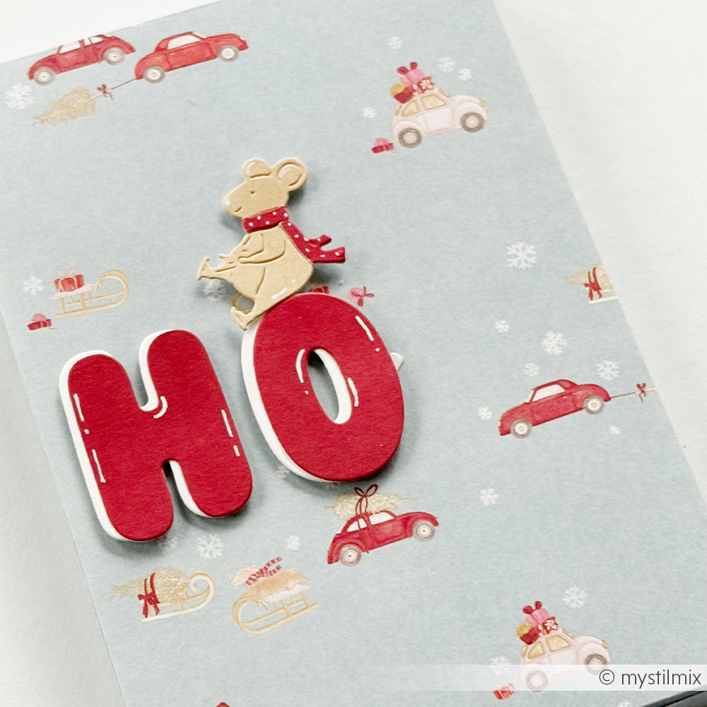 Eine Maus mit Schal sitzt auf einem roten Buchstaben O auf einem Designpapier mit Autos und Geschenken.