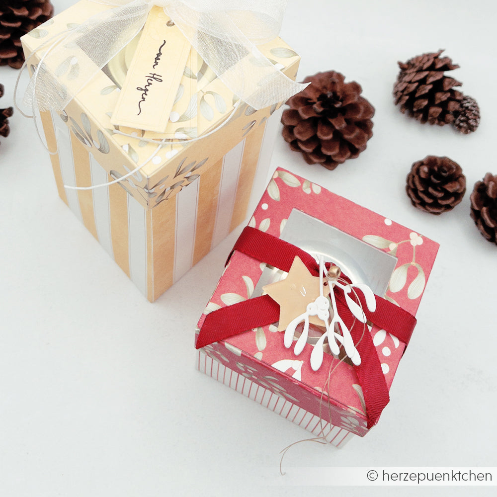 Zwei dekorative Geschenkboxen aus Designpapier mit Fenseldeckeln, Schleifen, Anhängern und Tannenzapfen.