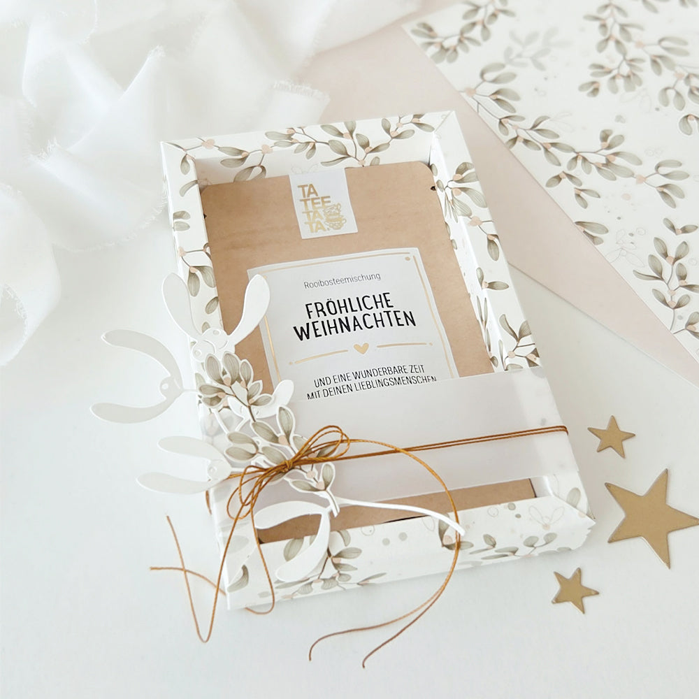 Eine Geschenkbox mit Rooibos-Tee, verpackt in festlichem Designpapier mit einem Etikett, auf dem „Fröhliche Weihnachten“ steht.