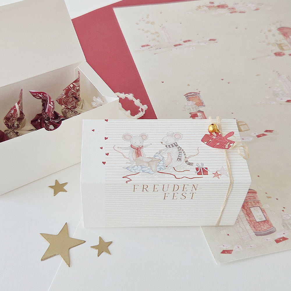 Eine weiße Geschenkbox mit einem „Weihnachtsmäuse“-Motiv, das zwei Mäuse zeigt, die mit der Aufschrift „Freuden Fest“ neben einer offenen Pralinenschachtel sitzen.