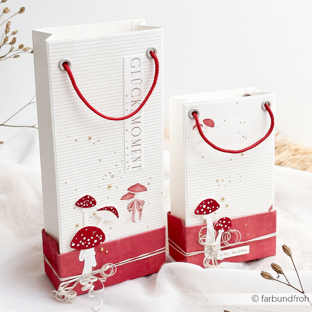 Zwei Projekte aus weißem Papier in Form von Geschenktüten, die mit roten Pilzillustrationen und roten Tragegriffen verziert sind.