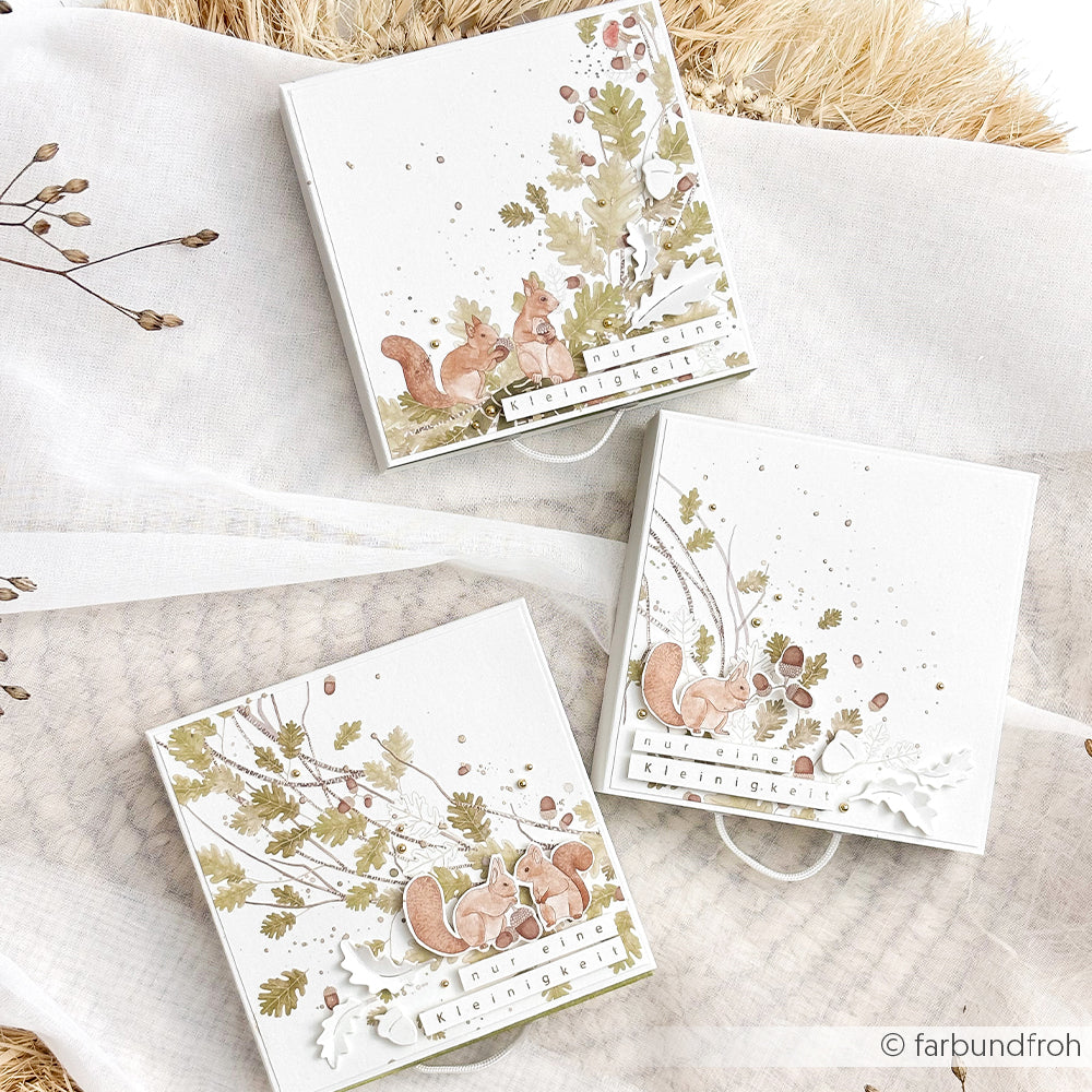 Drei quadratische Geschenkboxen, umhüllt von Designpapier mit Aquarellmotiven von Eichhörnchen, Eicheln und Blättern.