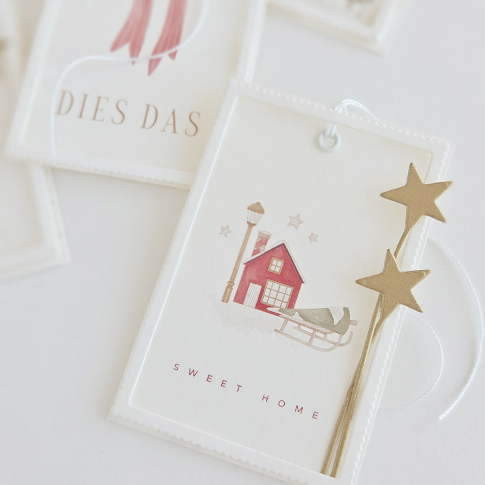 Geschenkanhänger aus der Papierkollektion „Wintermagie“ mit der Abbildung eines roten Hauses und eines Schlittens, verziert mit goldenen Sternen und dem Schriftzug „SWEET HOME“.