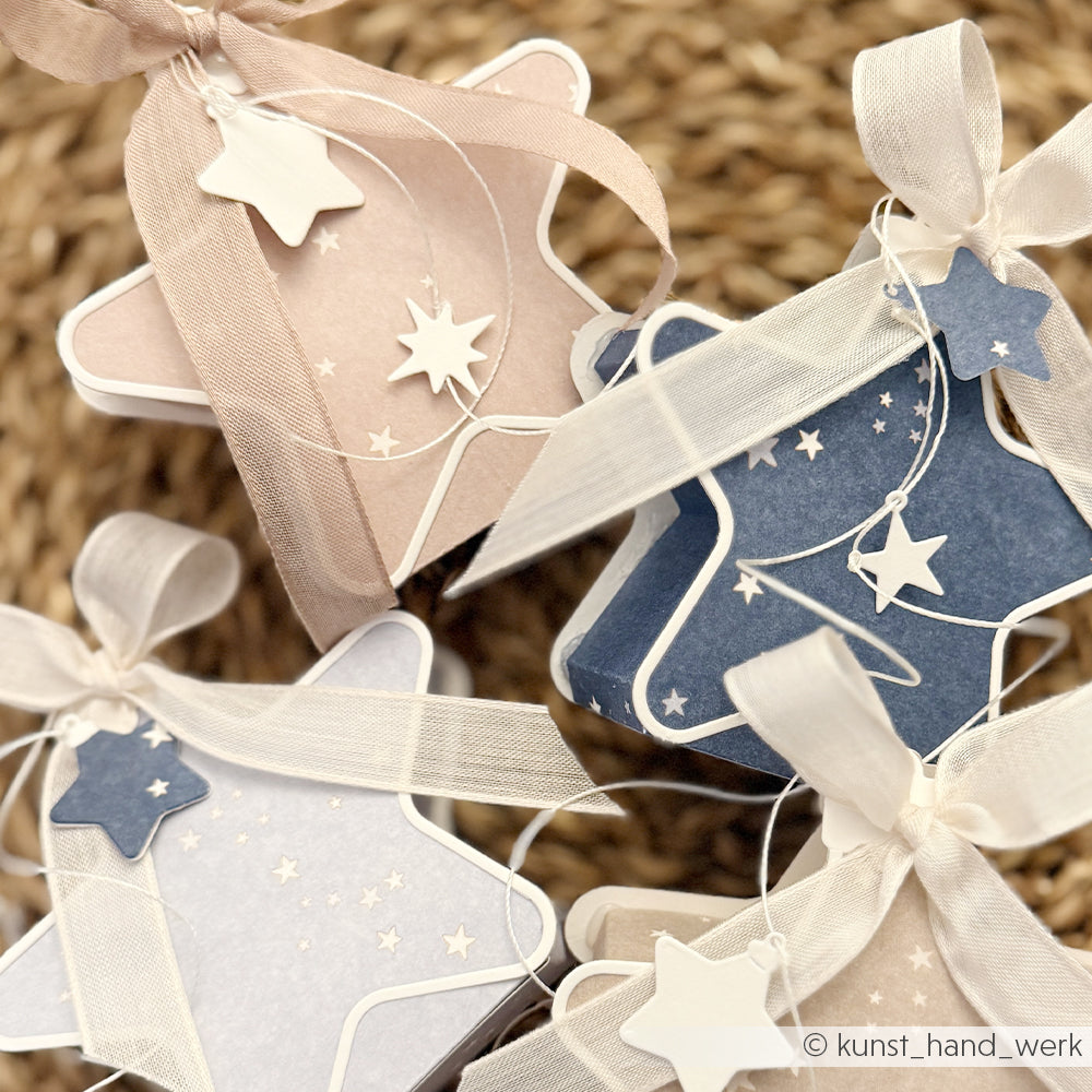 Vier sternförmige Geschenkboxen in Blau- und Beigetönen, aus Designpapier gefertigt und mit Bändern sowie kleinen Sternanhängern verziert.